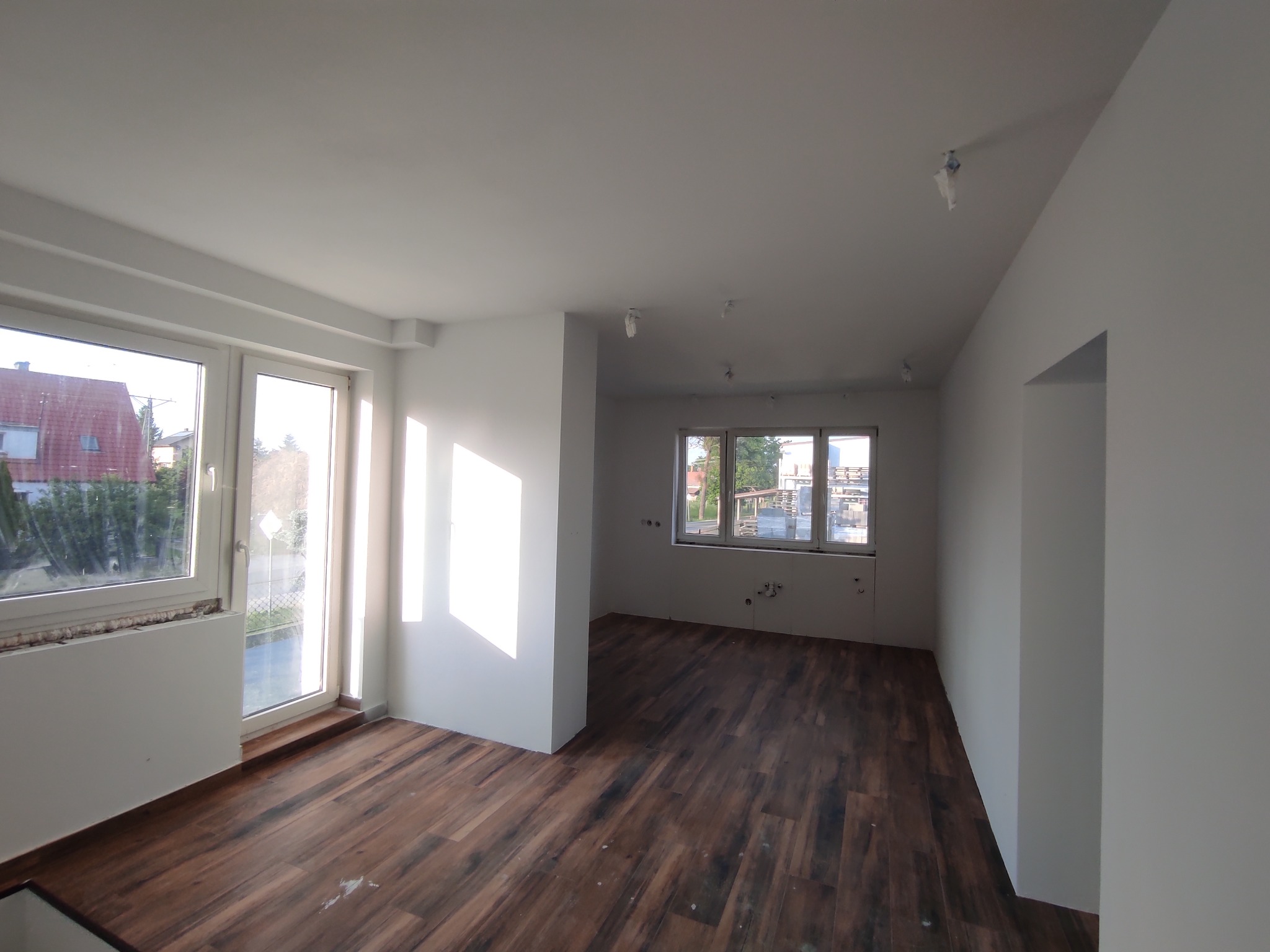 Jasne wnętrze po remoncie: świeżo pomalowane ściany, ciemne panele podłogowe, okna z widokiem na zieleń. Oświetlenie sufitowe w folii ochronnej. Minimalistyczny design.