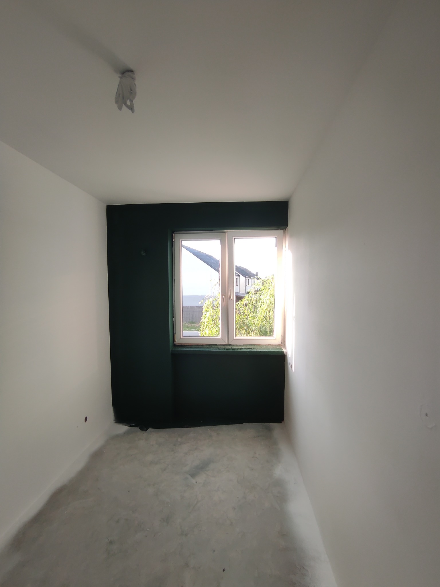 Pomalowany pokój na biało z ciemnozieloną ścianą z oknem. Na suficie widoczny element instalacji. Podłoga przygotowana do dalszych prac wykończeniowych.