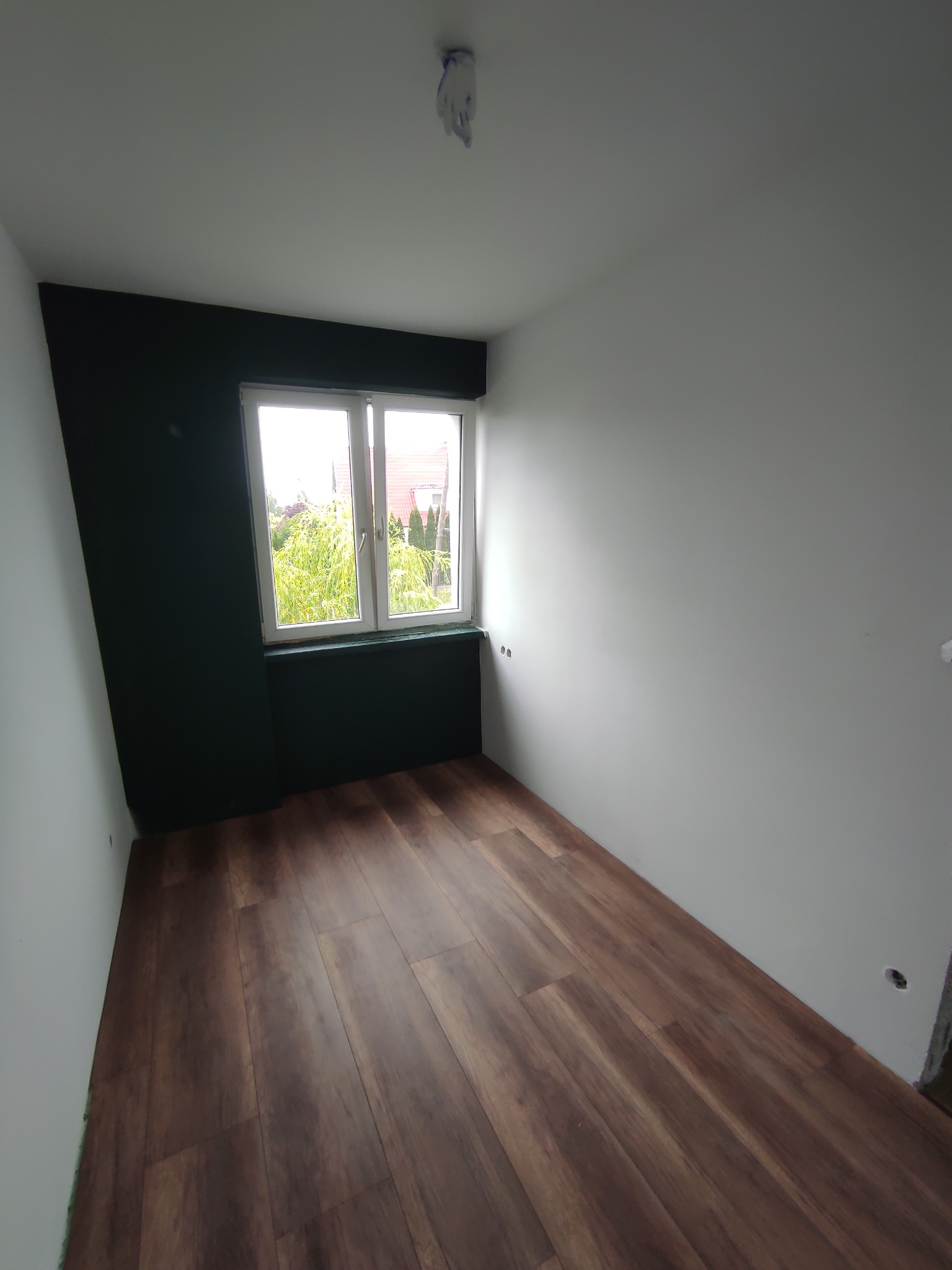 Pokój po remoncie: świeżo położony parkiet w odcieniach brązu, pomalowana na biało ściana z oknem i fragment ściany w kolorze ciemnej zieleni. Widok na zewnątrz.