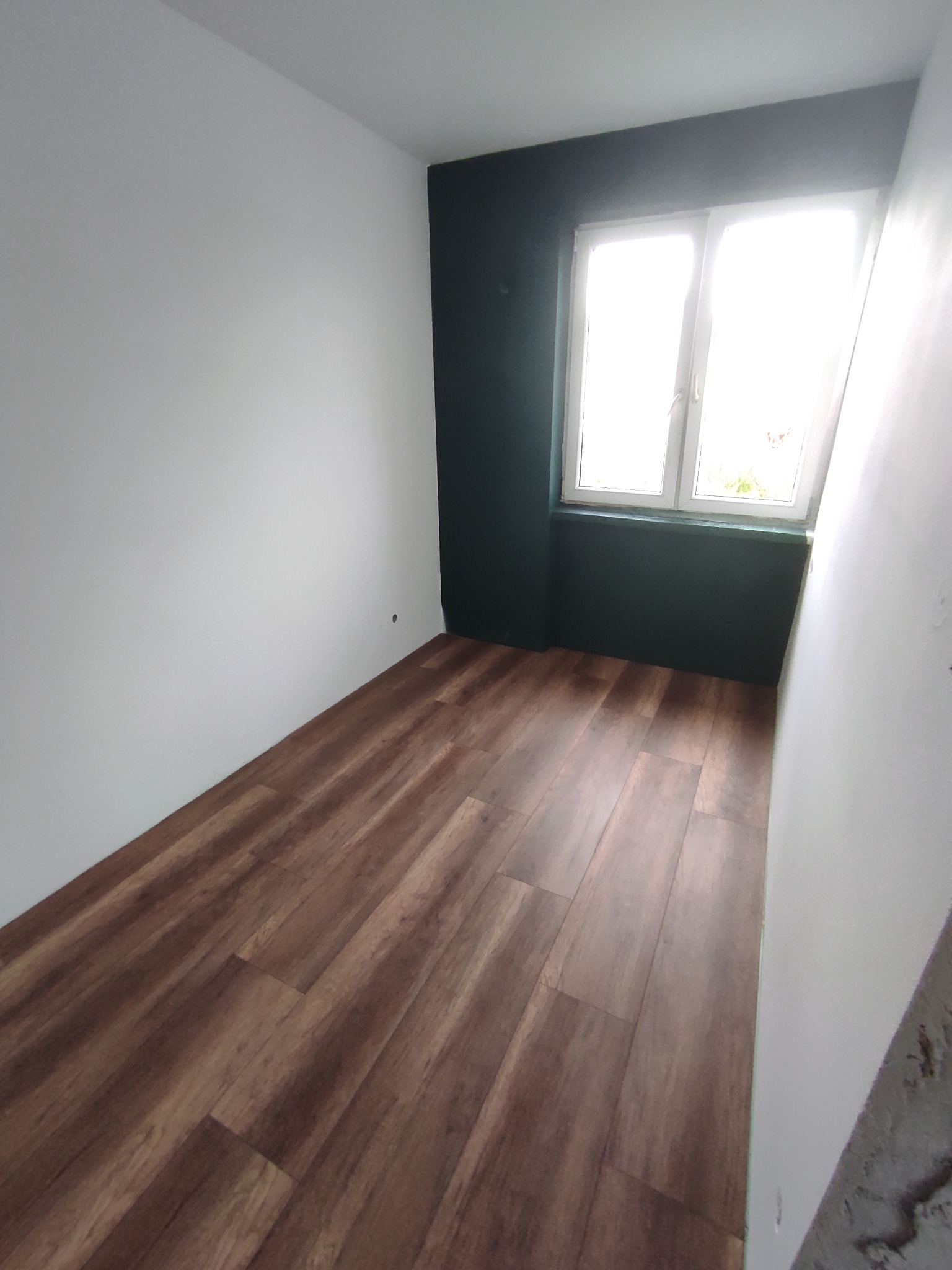 Pomieszczenie po remoncie: świeżo ułożony parkiet w odcieniach brązu, pomalowane ściany na biało i ciemnozielono. Okno z widokiem na zewnątrz. Jasne wnętrze.