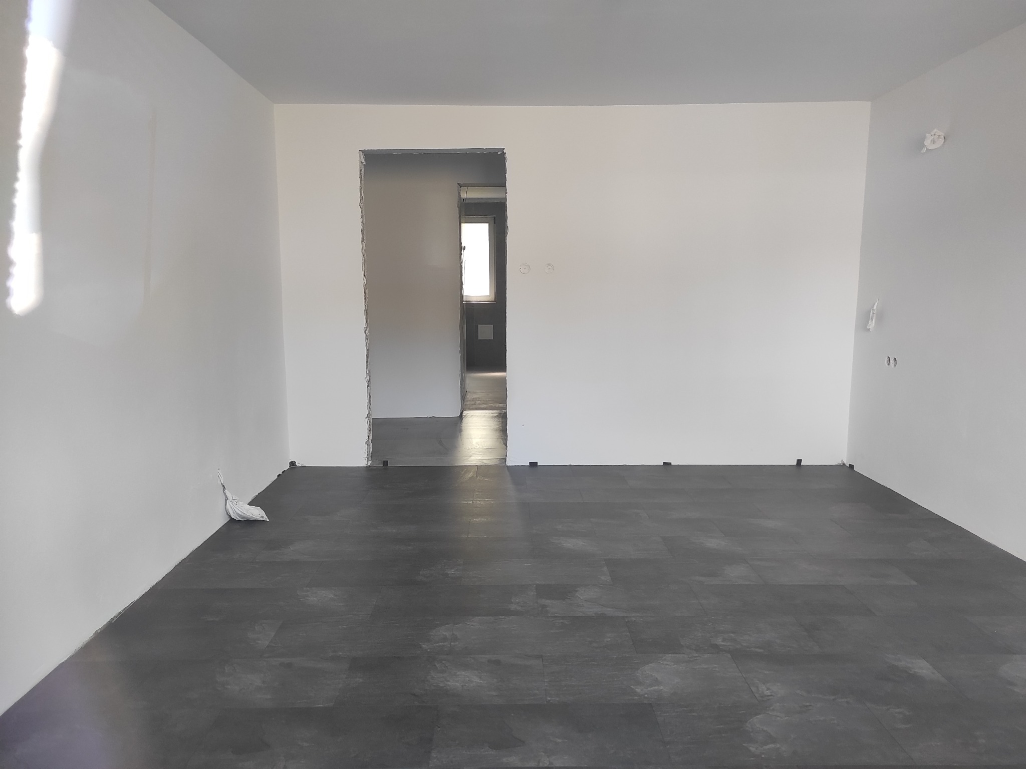 Ukończone układanie ciemnej podłogi w pustym, jasnym pokoju z widokiem na kolejne pomieszczenie przez otwór drzwiowy. Widoczny worek z resztkami materiałów.
