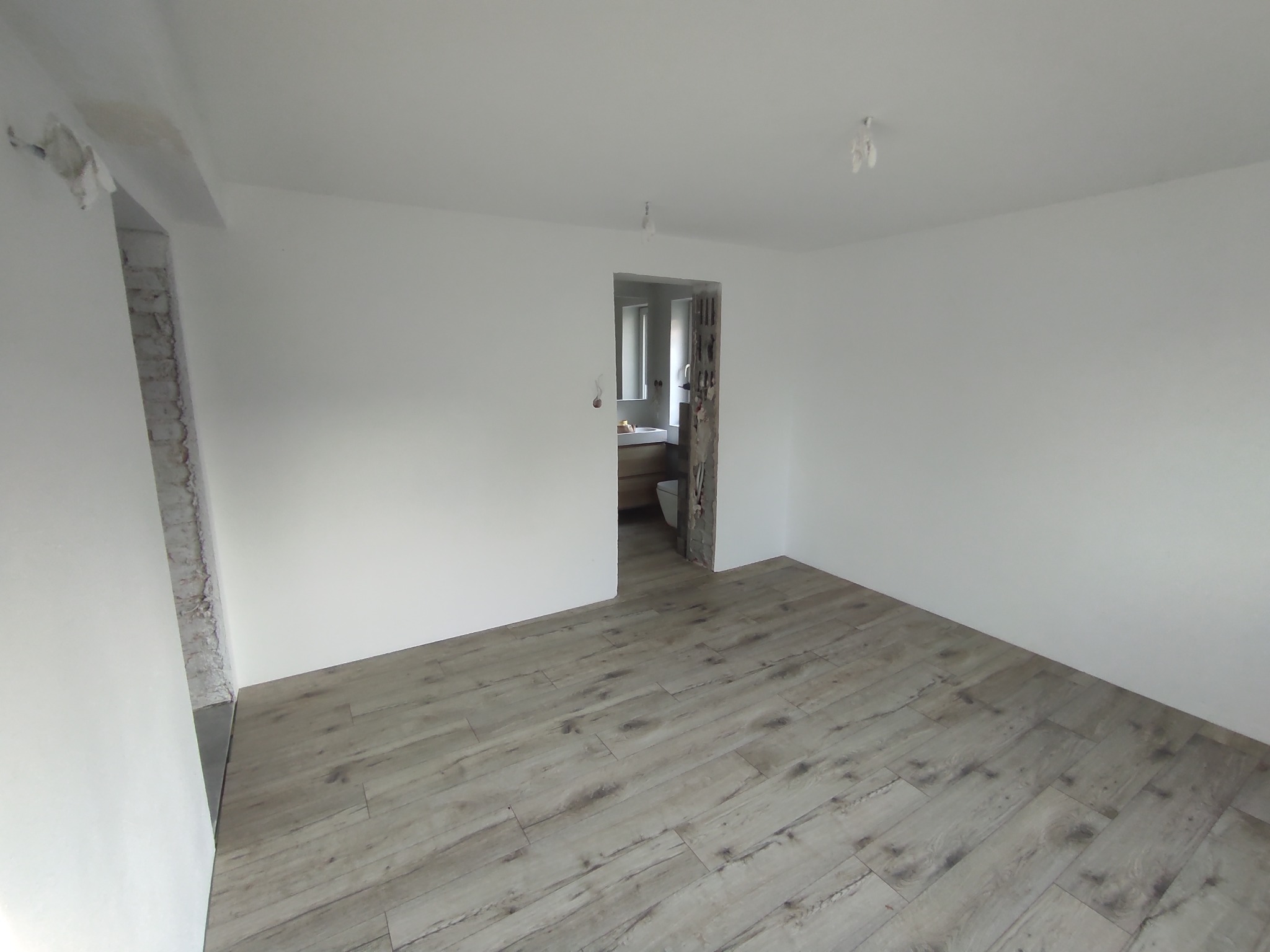 Jasne panele podłogowe ułożone w jodełkę w pokoju po remoncie. Ściany białe, gładkie, z fragmentami surowego muru przy przejściu do kolejnego pomieszczenia.