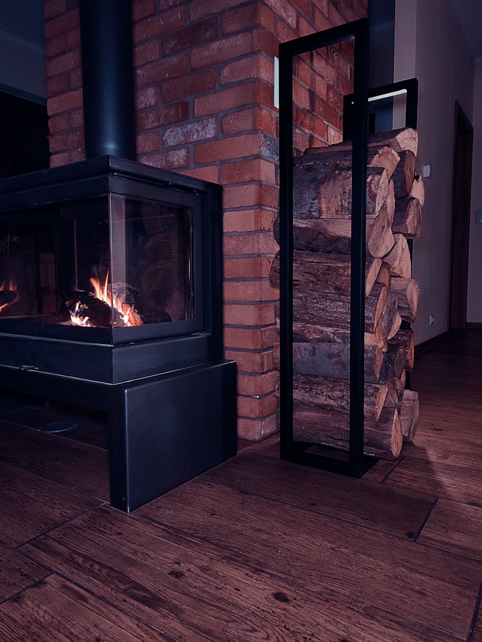 Nowoczesny kominek z paleniskiem i metalowym obramowaniem, ceglana ściana oraz stojak na drewno z czarną ramą na parkiecie.
