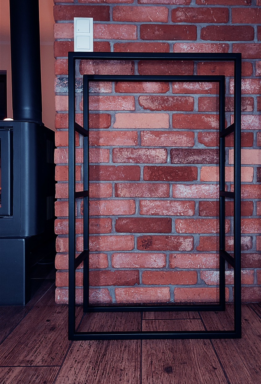 Czarna, metalowa konstrukcja regału na tle ceglanej ściany i drewnianej podłogi, widoczny fragment pieca.