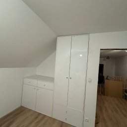 Bartek Prestigio - Biała szafa wnękowa z połyskiem, dopasowana do skosu dachu, z widocznymi gniazdkami elektrycznymi i fragmentem pokoju dziecięcego w tle.