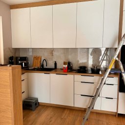 Biała kuchnia z drewnianymi akcentami i marmurowym backsplashiem, widoczna drabina sugeruje montaż lub wykończenie. Nowoczesny design z czarnymi uchwytami i armaturą.