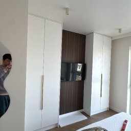 Białe szafy wnękowe z długimi, złotymi uchwytami, wkomponowane w brązową ścianę z telewizorem. Na podłodze parkiet w jodełkę. Widoczny fragment drabiny malarskiej.