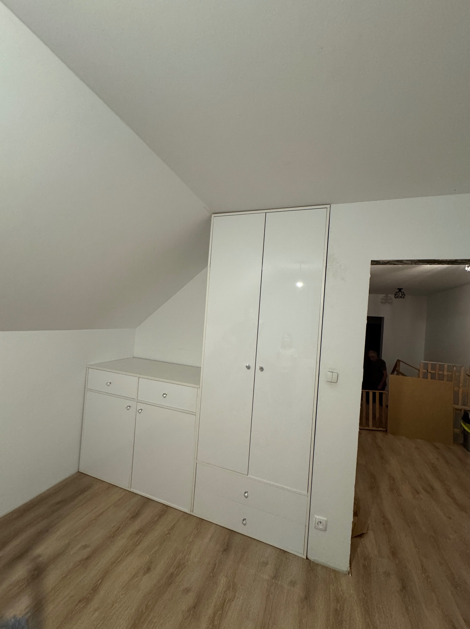 Biała szafa wnękowa z połyskiem, dopasowana do skosu dachu, z widocznymi gniazdkami elektrycznymi i fragmentem pokoju dziecięcego w tle.