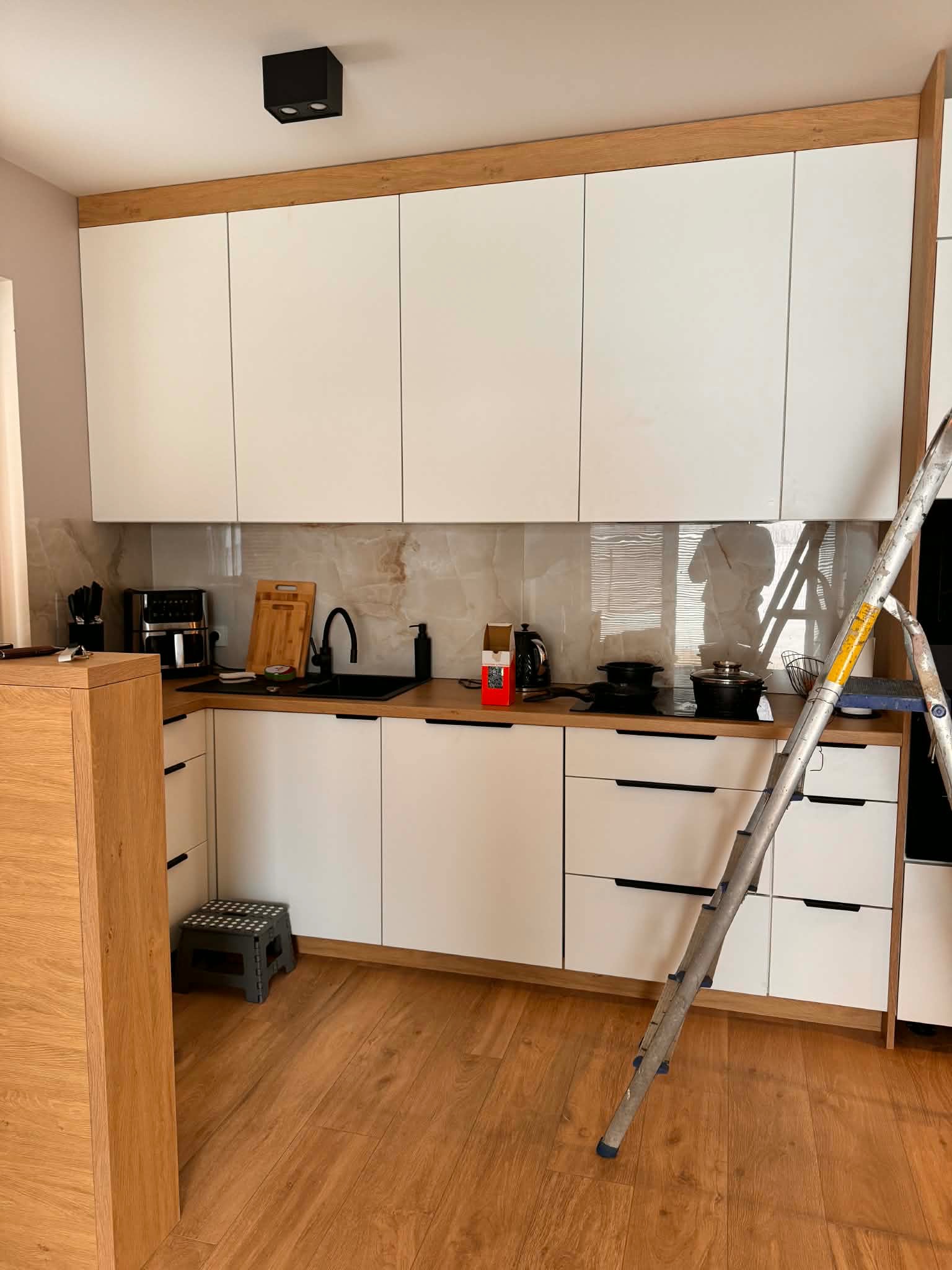 Biała kuchnia z drewnianymi akcentami i marmurowym backsplashiem, widoczna drabina sugeruje montaż lub wykończenie. Nowoczesny design z czarnymi uchwytami i armaturą.