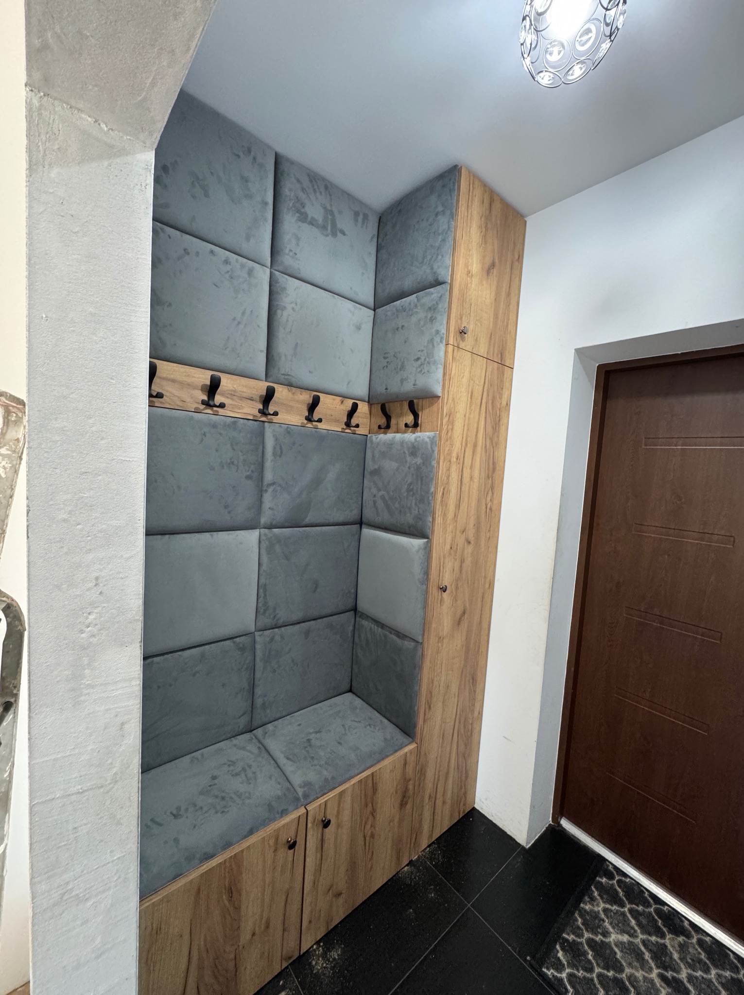 Zabudowa wnęki z siedziskiem i wieszakami, wykończona drewnopodobną płytą i tapicerowanymi panelami w odcieniach szarości. Funkcjonalne rozwiązanie do przedpokoju.