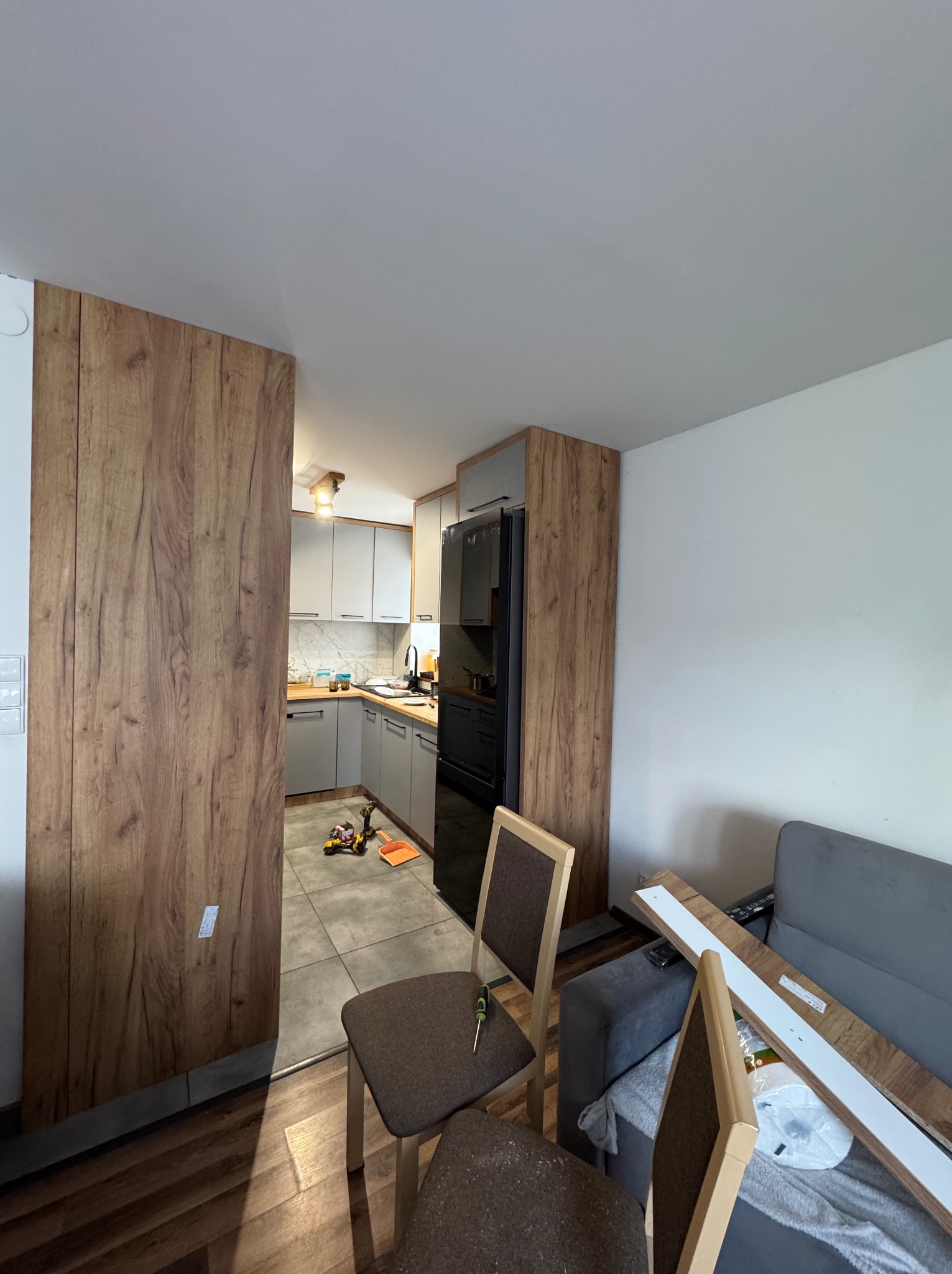 Montaż kuchni: widok na szarą zabudowę z czarną lodówką, drewniane panele na ścianie i narzędzia na podłodze. Częściowo widoczne krzesła i sofa.