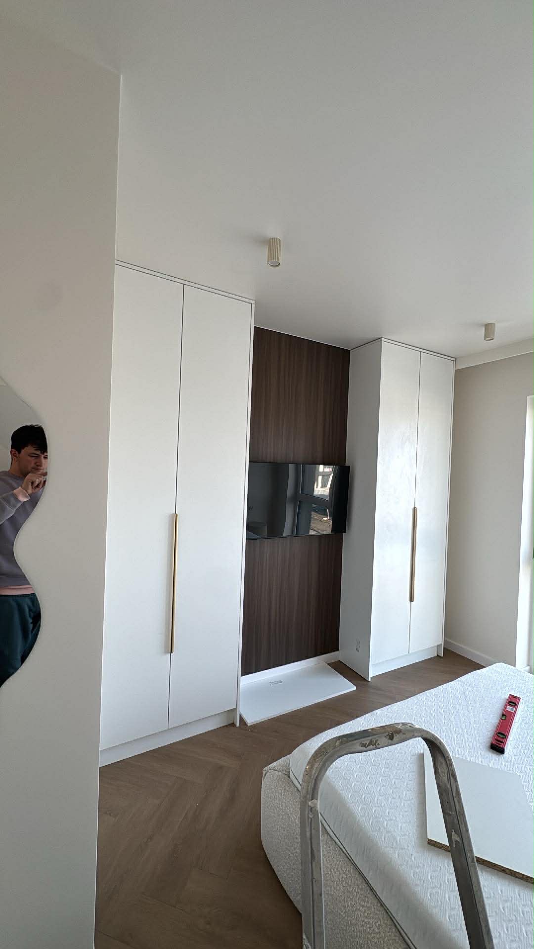 Białe szafy wnękowe z długimi, złotymi uchwytami, wkomponowane w brązową ścianę z telewizorem. Na podłodze parkiet w jodełkę. Widoczny fragment drabiny malarskiej.