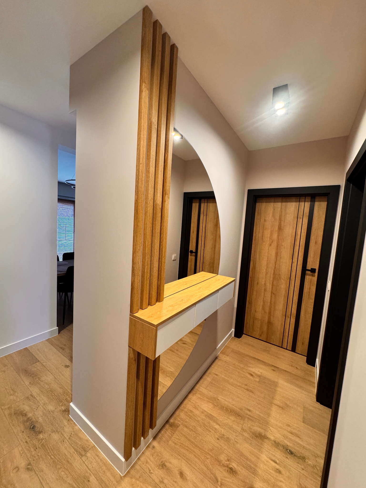 Nowoczesny przedpokój z lustrem w kształcie łuku, drewnianymi lamelami i blatem w kolorze naturalnego drewna. Na podłodze panele w odcieniach brązu, drzwi z czarną ramą.