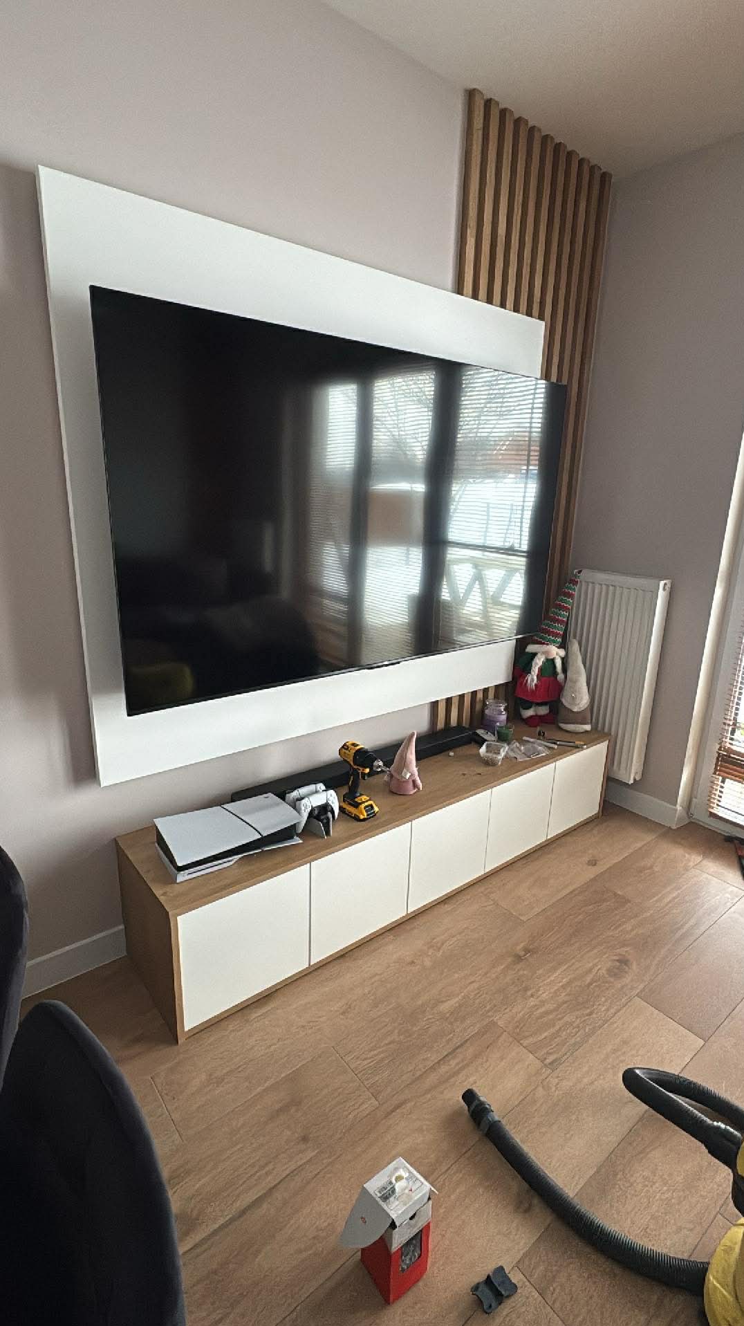 Nowoczesna szafka RTV z białymi frontami i drewnianym blatem, na niej konsola do gier, wiertarka i dekoracje. Na ścianie duży telewizor w białej ramie i drewniane lamele.