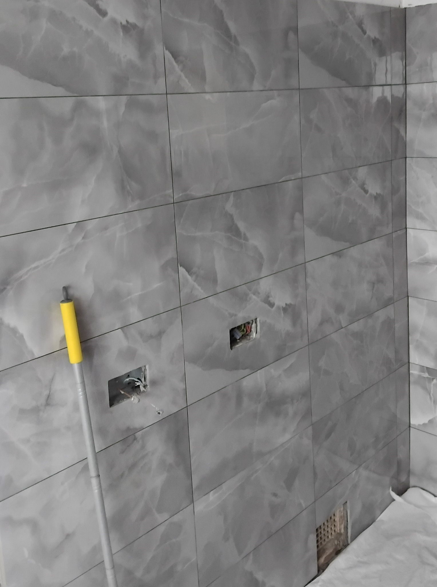 Szare, marmurowe płytki ścienne w łazience, z widocznymi puszkami elektrycznymi i fragmentem narzędzia z żółtą rączką w rogu. Układ poziomy, fuga wąska.