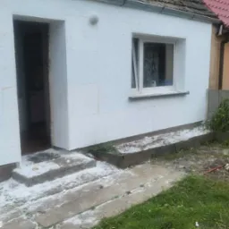 Dom w trakcie ocieplania: biały styropian na ścianach, widoczne wejście i okno. Fragment dachu z dachówką w tle. Ślady styropianu na schodach i trawie.