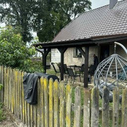 Termo - Tech Termomodernizacja Kamil Żurawowicz - Dom z brązowym dachem widoczny zza drewnianego płotu z porostami. Zadaszony taras z meblami ogrodowymi. Widoczny fragment elewacji i osoba na tarasie.