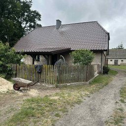 Termo - Tech Termomodernizacja Kamil Żurawowicz - Dom z nowym, brązowym dachem z dachówki, widoczny z boku, z drewnianym ogrodzeniem, taczką i stertą piasku. Na drugim planie budynek gospodarczy.