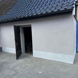 Termo - Tech Termomodernizacja Kamil Żurawowicz - Wykończona elewacja budynku z ciemną dachówką, otwartymi drewnianymi drzwiami i szarą kostką brukową. Widoczny fragment podbitki i okno dachowe.