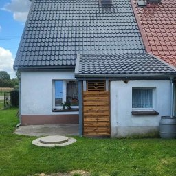 Termo - Tech Termomodernizacja Kamil Żurawowicz - Dom jednorodzinny z nowym, szarym dachem z dachówki ceramicznej, kontrastującym z czerwoną dachówką na dobudówce. Widoczna drewniana osłona i fragment elewacji.