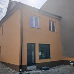 Dwukondygnacyjny dom z elewacją w odcieniach beżu i brązu, z ciemnymi oknami i drzwiami. Widoczny fragment dachu i rynny. Mały budynek sąsiaduje z większym.