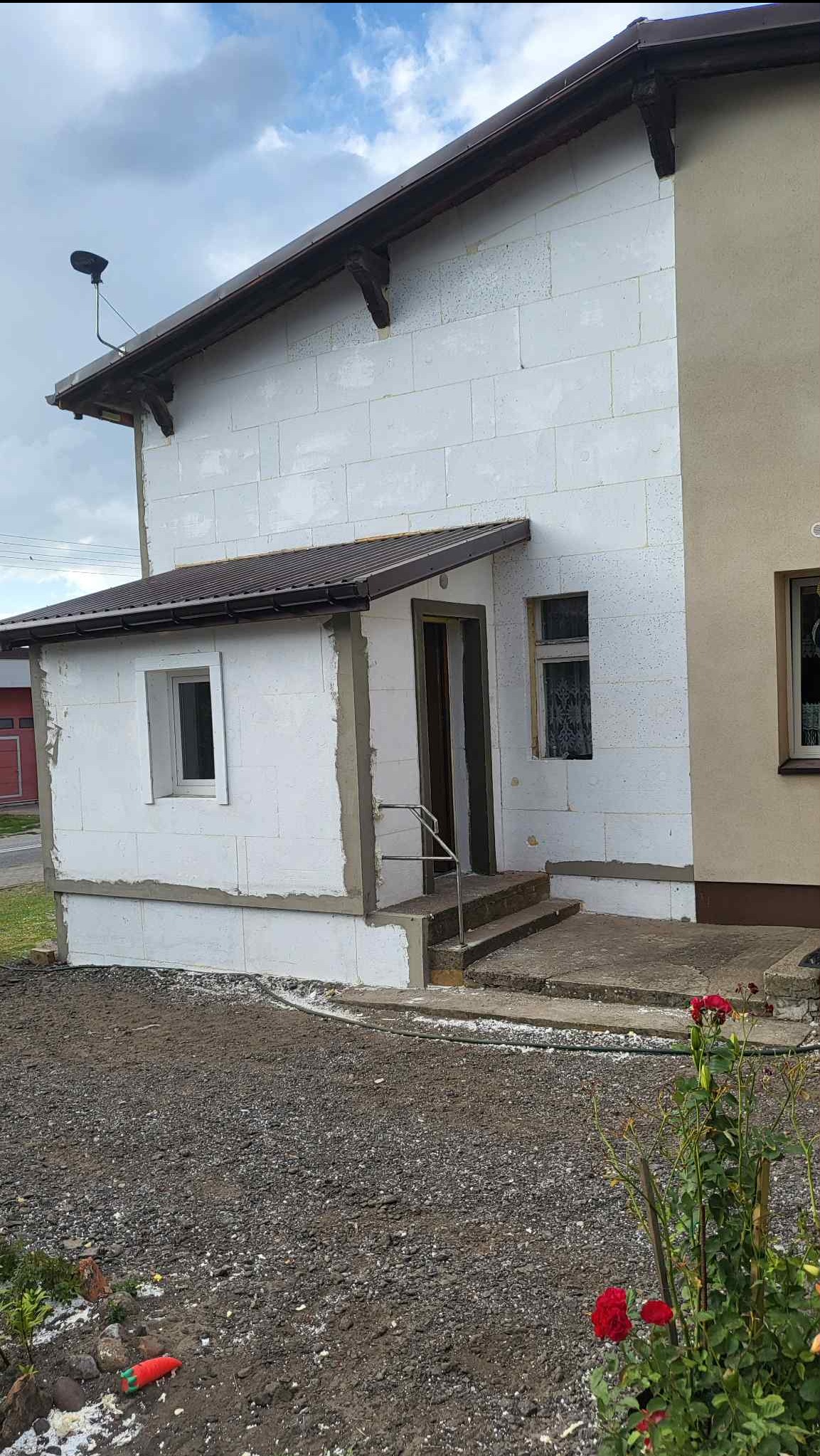 Dom w trakcie ocieplania styropianem, widoczne płyty i przygotowane wejście z zadaszeniem. Część elewacji otynkowana. W tle pochmurne niebo.