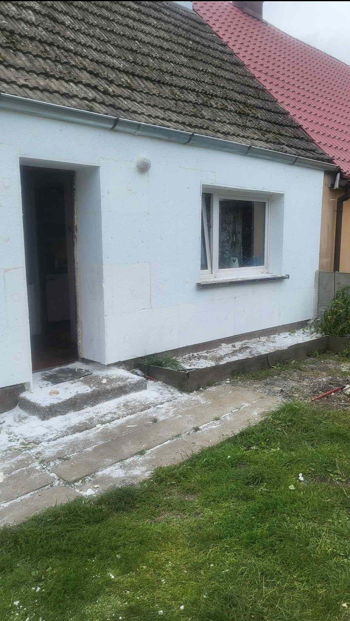 Dom w trakcie ocieplania: biały styropian na ścianach, widoczne wejście i okno. Fragment dachu z dachówką w tle. Ślady styropianu na schodach i trawie.
