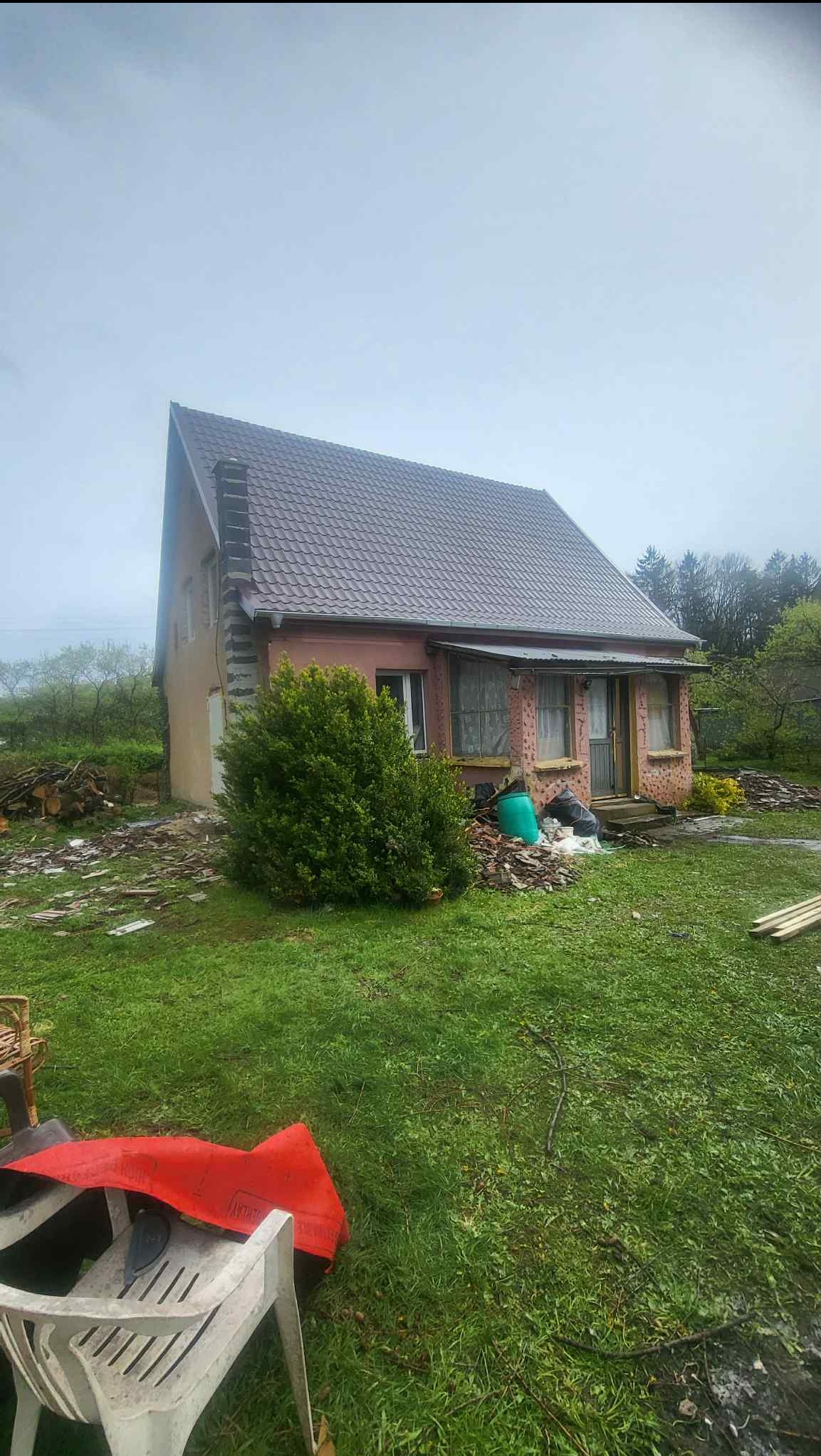 Niski dom z brązowym dachem po renowacji, otoczony zielenią i gruzem. Widoczne fragmenty elewacji i nowy dach. Niebo pochmurne.