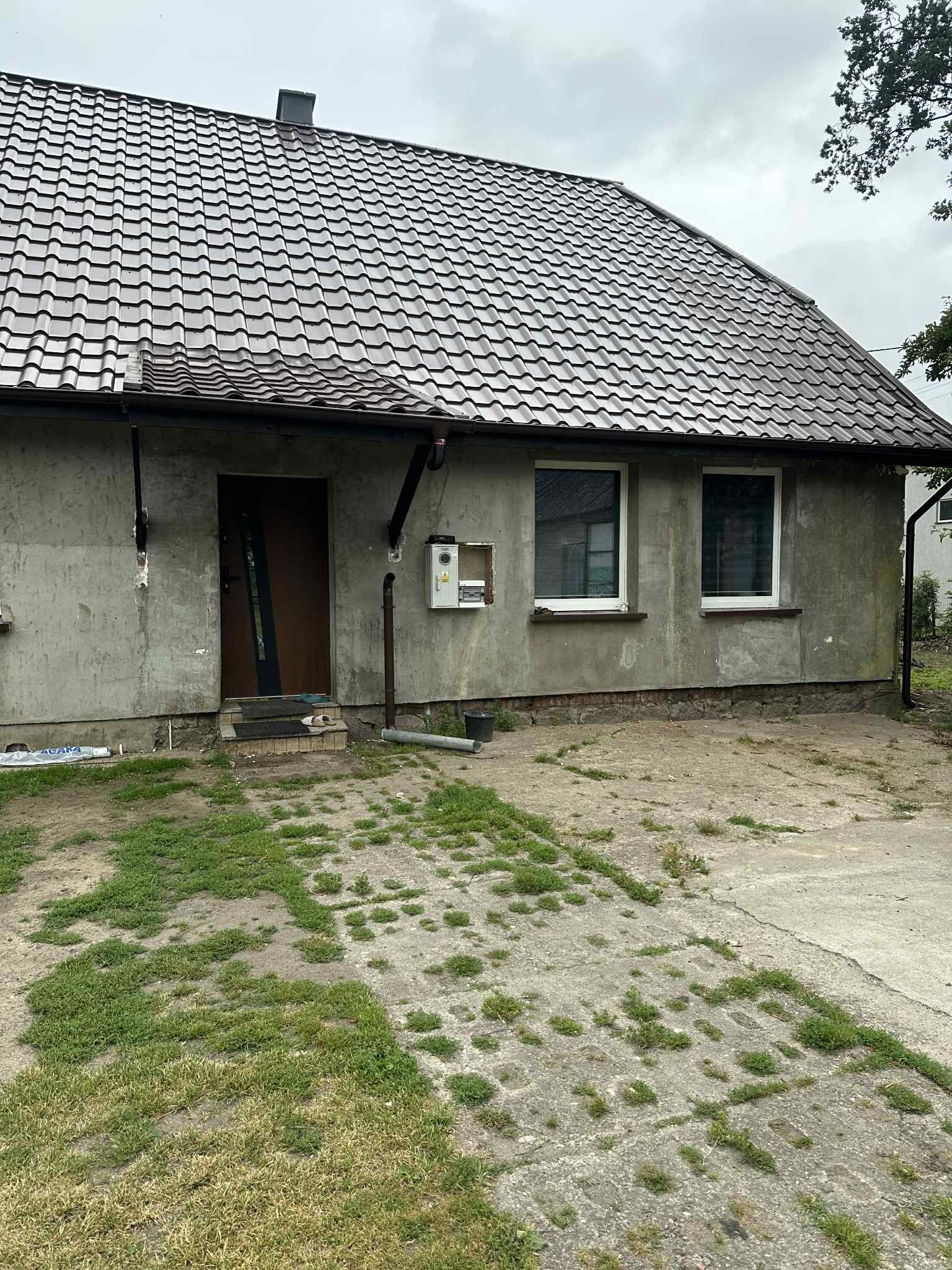 Dom z brązowym dachem z dachówki karpiówki i surową elewacją, widoczny fragment betonowego podjazdu porośniętego trawą. Widok na budynek w pochmurny dzień.