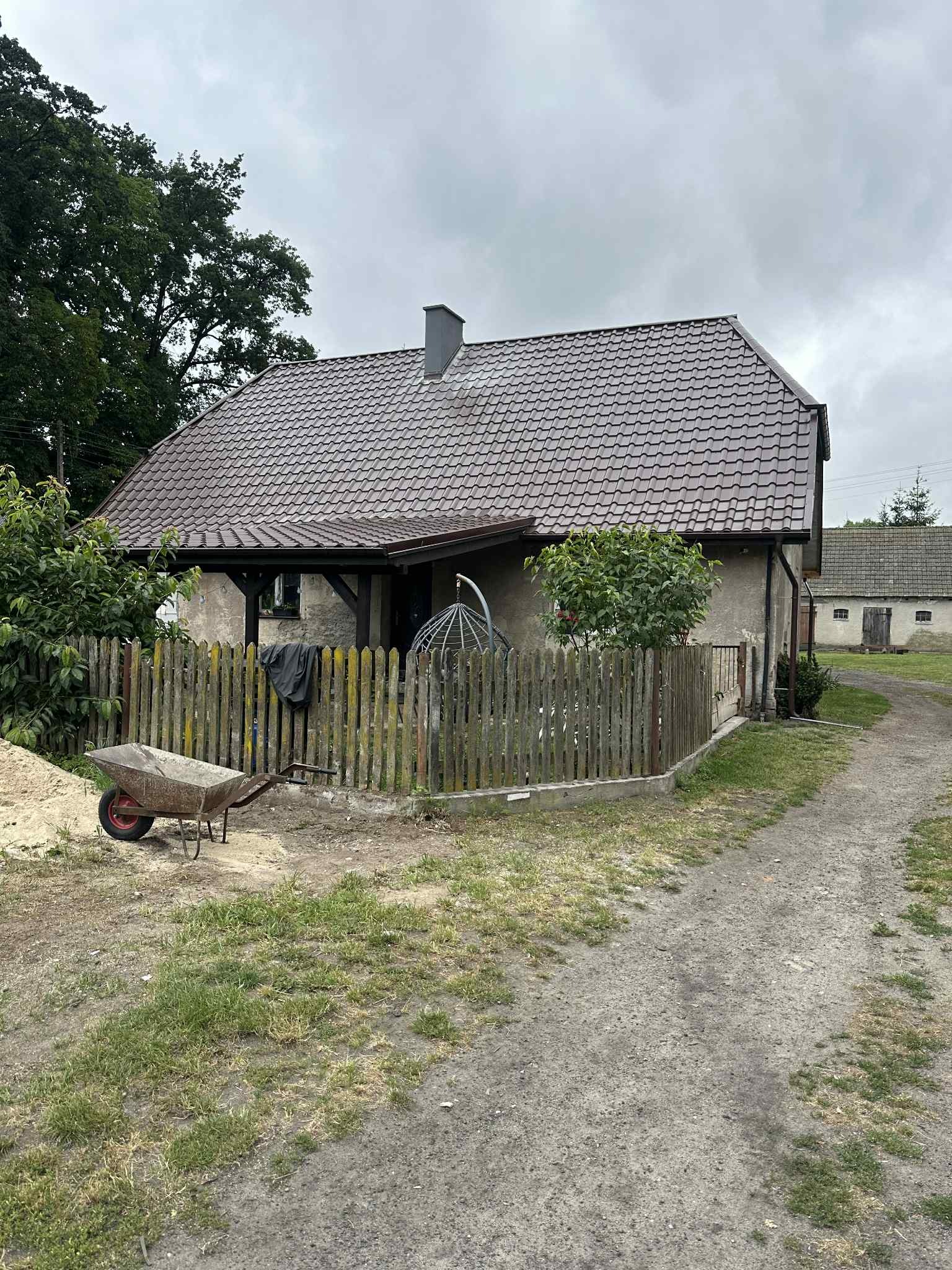 Dom z nowym, brązowym dachem z dachówki, widoczny z boku, z drewnianym ogrodzeniem, taczką i stertą piasku. Na drugim planie budynek gospodarczy.