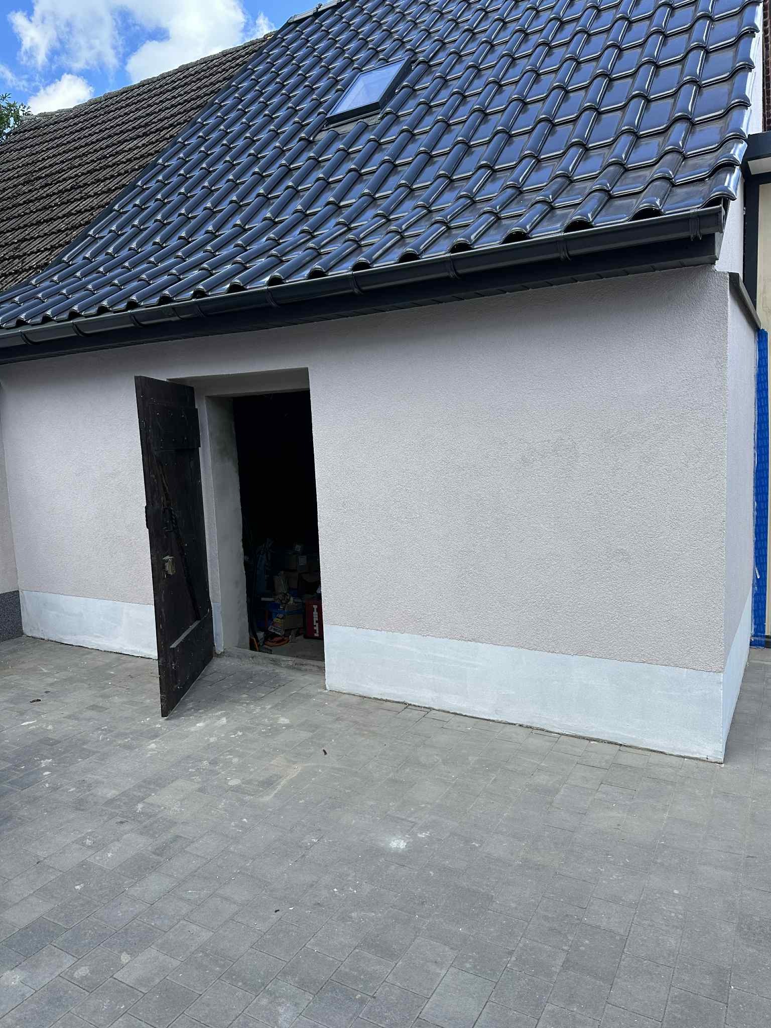 Wykończona elewacja budynku z ciemną dachówką, otwartymi drewnianymi drzwiami i szarą kostką brukową. Widoczny fragment podbitki i okno dachowe.