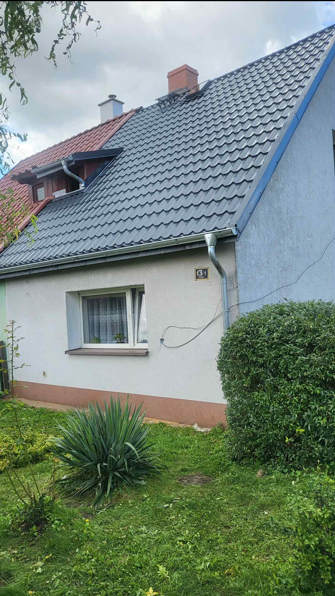 Dom z nowym, ciemnoszarym dachem z blachodachówki trapezowej i orynnowaniem. Widoczna elewacja budynku z numerem 13/1.