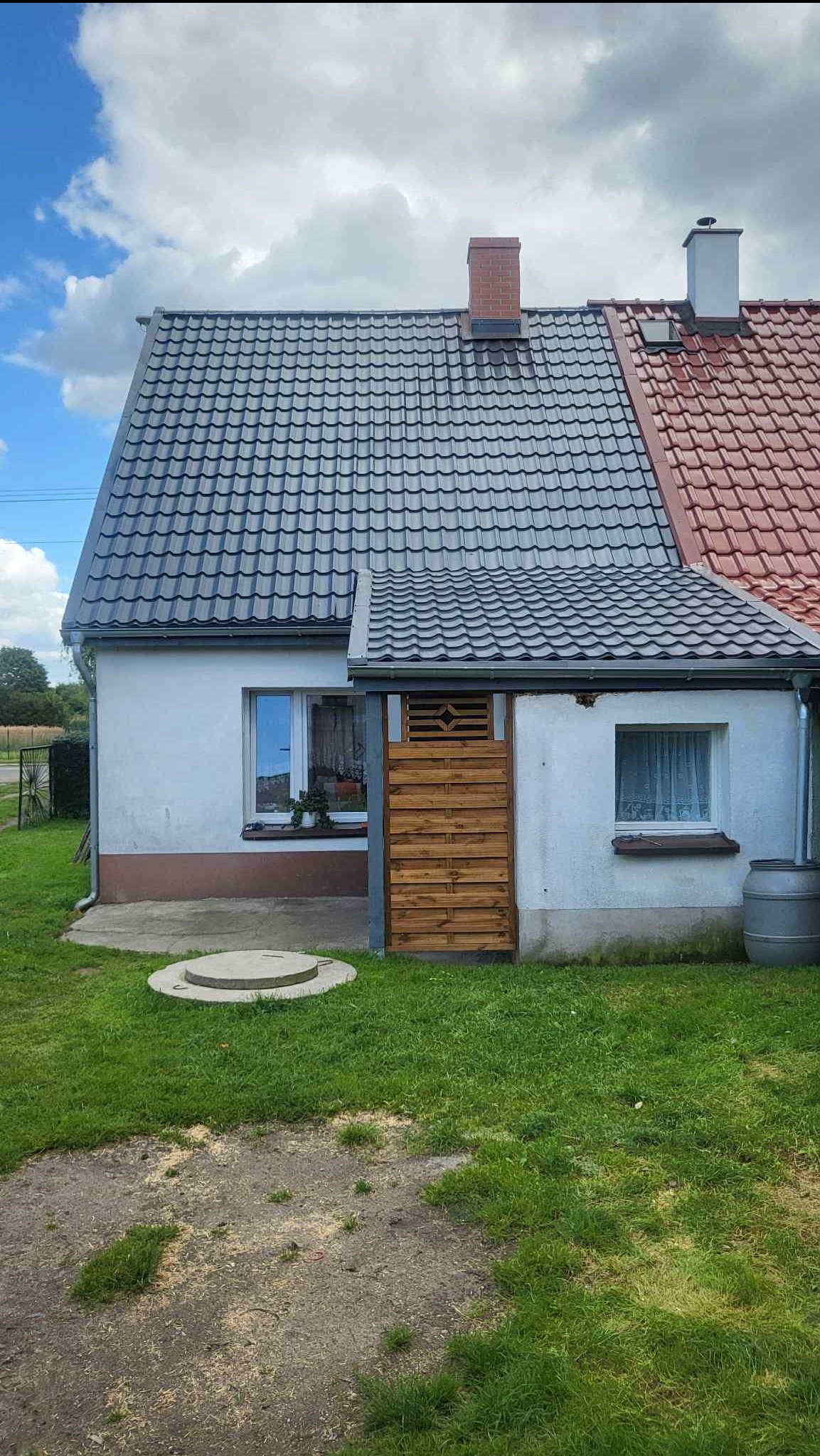 Dom jednorodzinny z nowym, szarym dachem z dachówki ceramicznej, kontrastującym z czerwoną dachówką na dobudówce. Widoczna drewniana osłona i fragment elewacji.