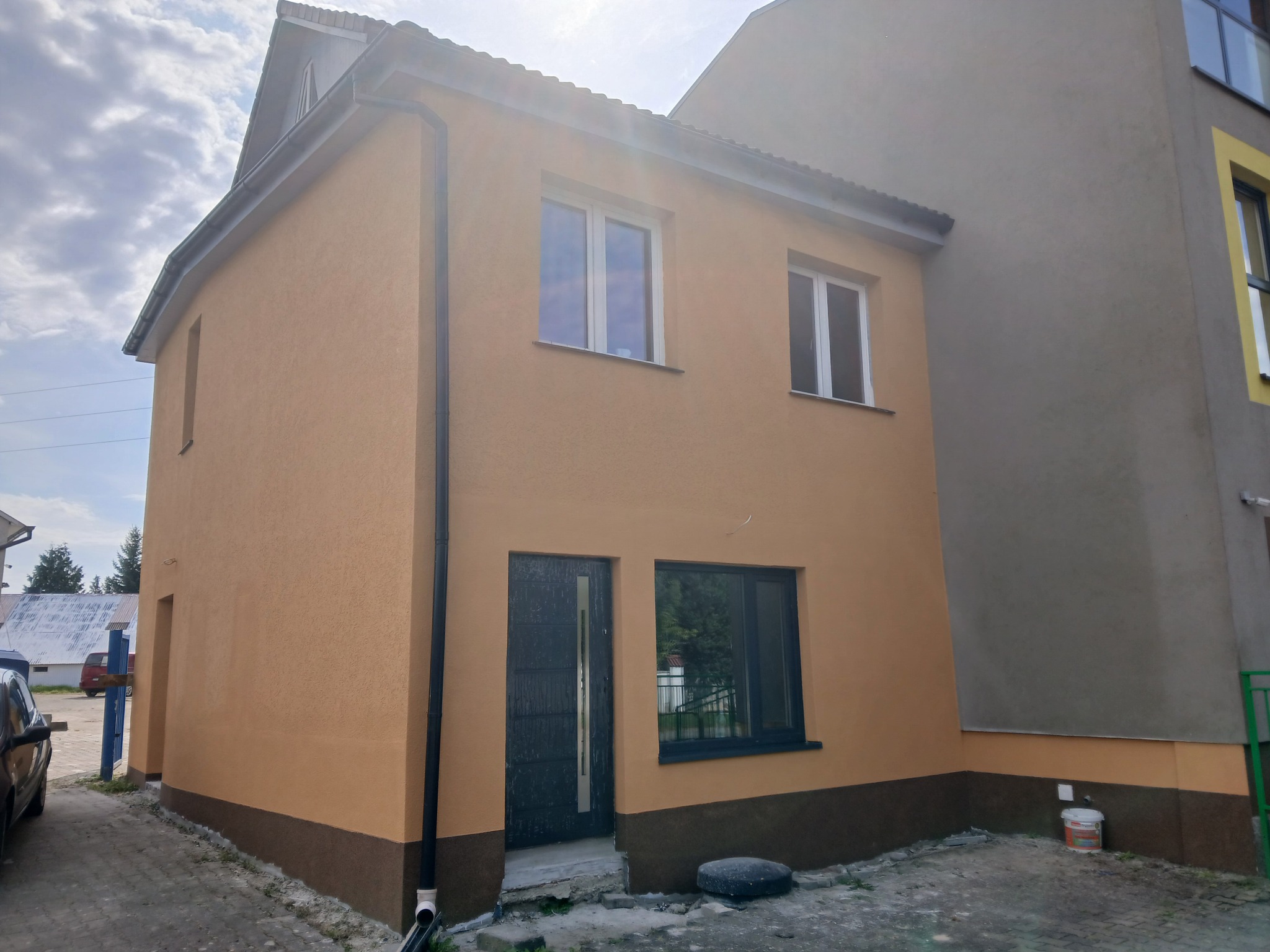 Dwukondygnacyjny dom z elewacją w odcieniach beżu i brązu, z ciemnymi oknami i drzwiami. Widoczny fragment dachu i rynny. Mały budynek sąsiaduje z większym.