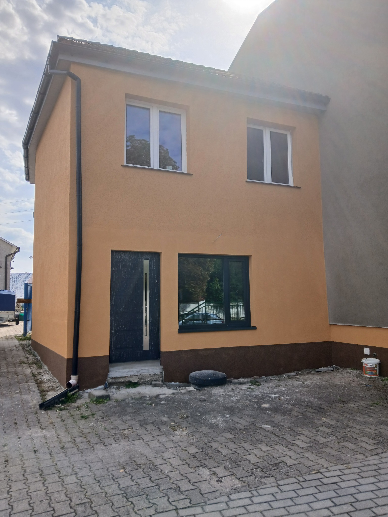 Dwukondygnacyjny dom z elewacją w odcieniach beżu i brązu, widoczne okna z białymi ramami i ciemne drzwi wejściowe. Dom po renowacji, z nową elewacją i orynnowaniem.