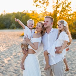 Foto Studio Izabela Godlewska - Rodzinna sesja w Warszawie: Mama trzyma synka wskazującego palcem w górę, tata córeczkę, wszyscy uśmiechnięci na tle piaszczystej plaży i drzew w ciepłym świetle.