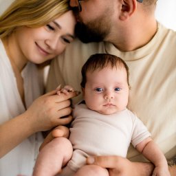 Foto Studio Izabela Godlewska - Rodzinne ujęcie: ojciec w okularach całuje matkę w czoło, trzymając na rękach niemowlę z intensywnym spojrzeniem, w jasnym wnętrzu.