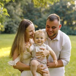 Foto Studio Izabela Godlewska - Uśmiechnięta rodzina w parku: mama w kwiecistej sukience trzyma synka, tata obejmuje ich. Delikatne światło podkreśla radosną atmosferę sesji rodzinnej.