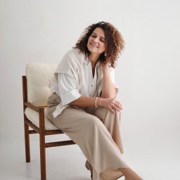 Foto Studio Izabela Godlewska - Kobieta z kręconymi włosami siedzi na krześle w jasnym wnętrzu, ubrana w beżowe spodnie i białą koszulę. Uśmiech na twarzy, delikatna biżuteria.