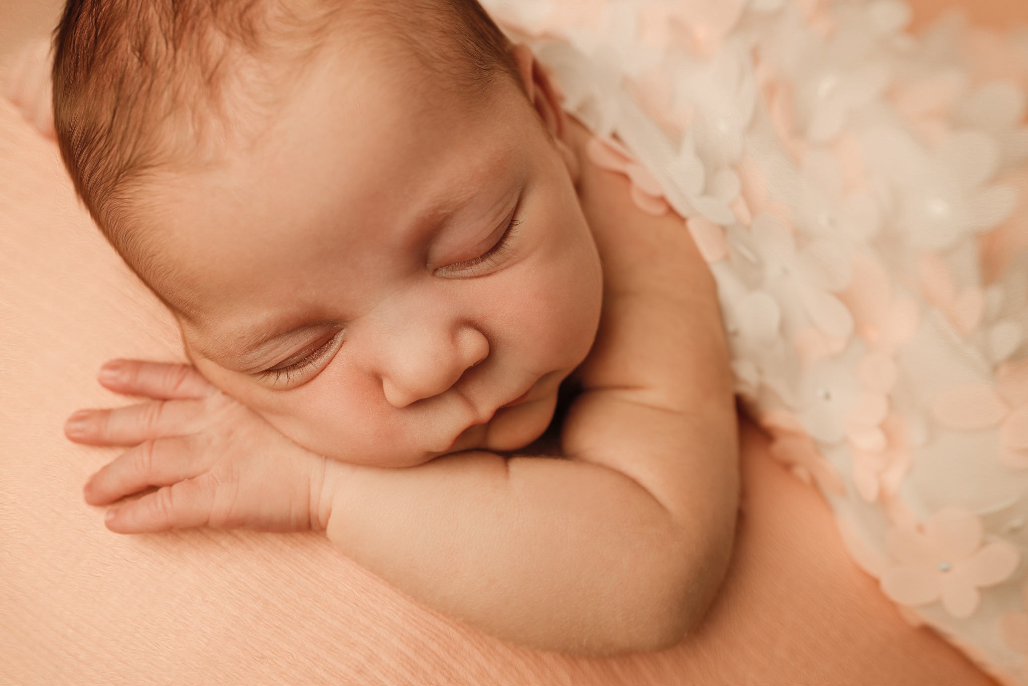 Śpiący noworodek z drobną rączką, na delikatnym, kremowym tle z kwiatową dekoracją. Zamknięte oczy, spokojny wyraz twarzy, miękkie światło.