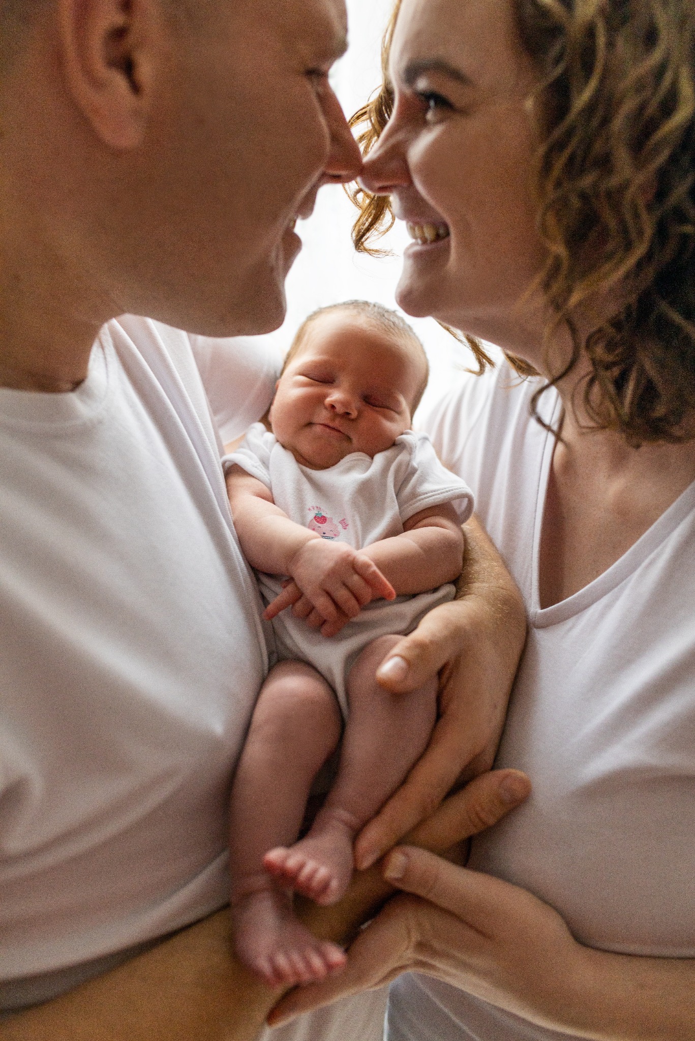 Rodzinne ujęcie: rodzice, ubrani w białe koszulki, trzymają na rękach śpiącego noworodka w białym body z aplikacją, zbliżając się do siebie nosami.