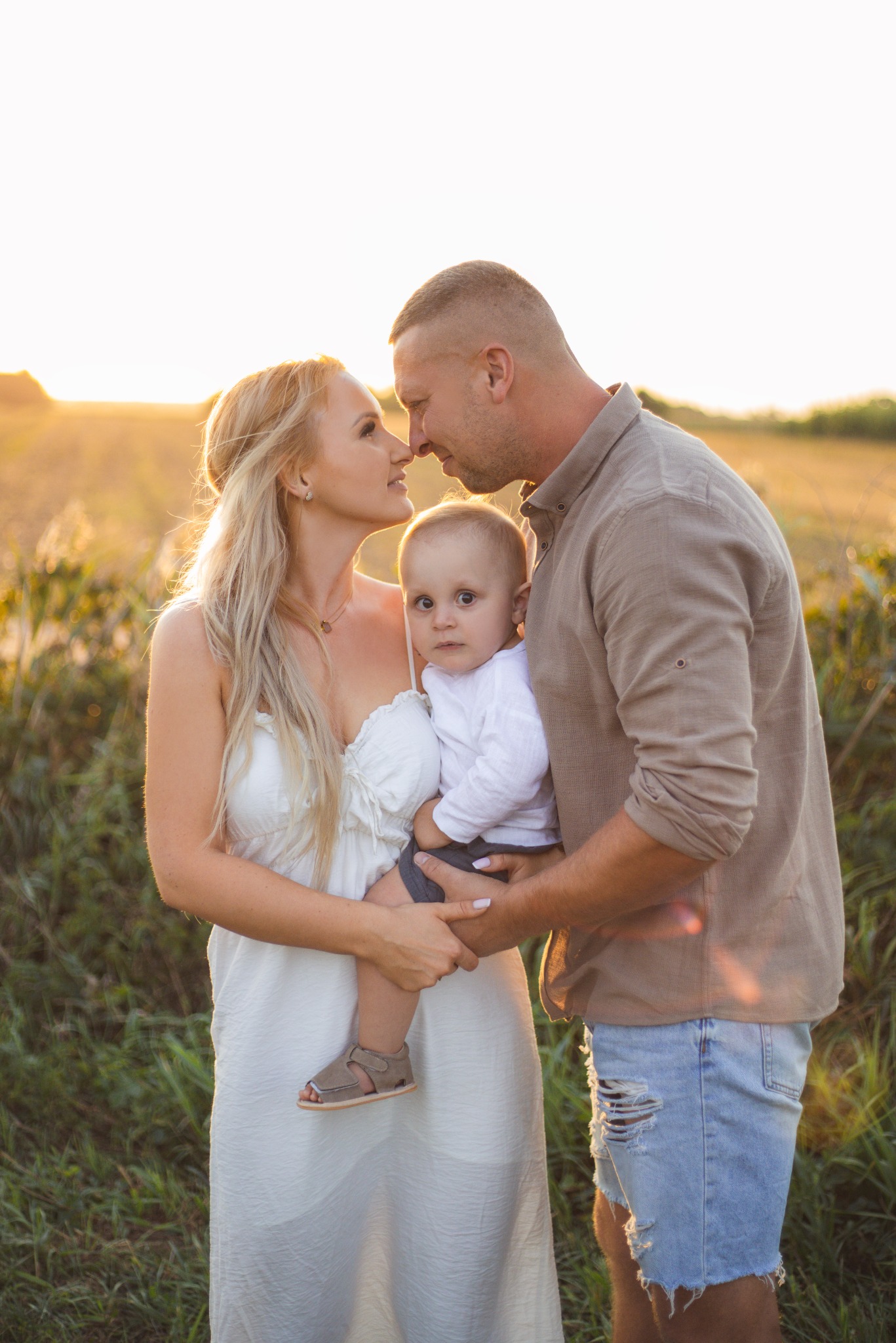 Uśmiechnięta rodzina: mama w białej sukience trzyma dziecko w koszuli, tata w koszuli i jeansowych spodenkach, na tle pola w promieniach zachodzącego słońca.