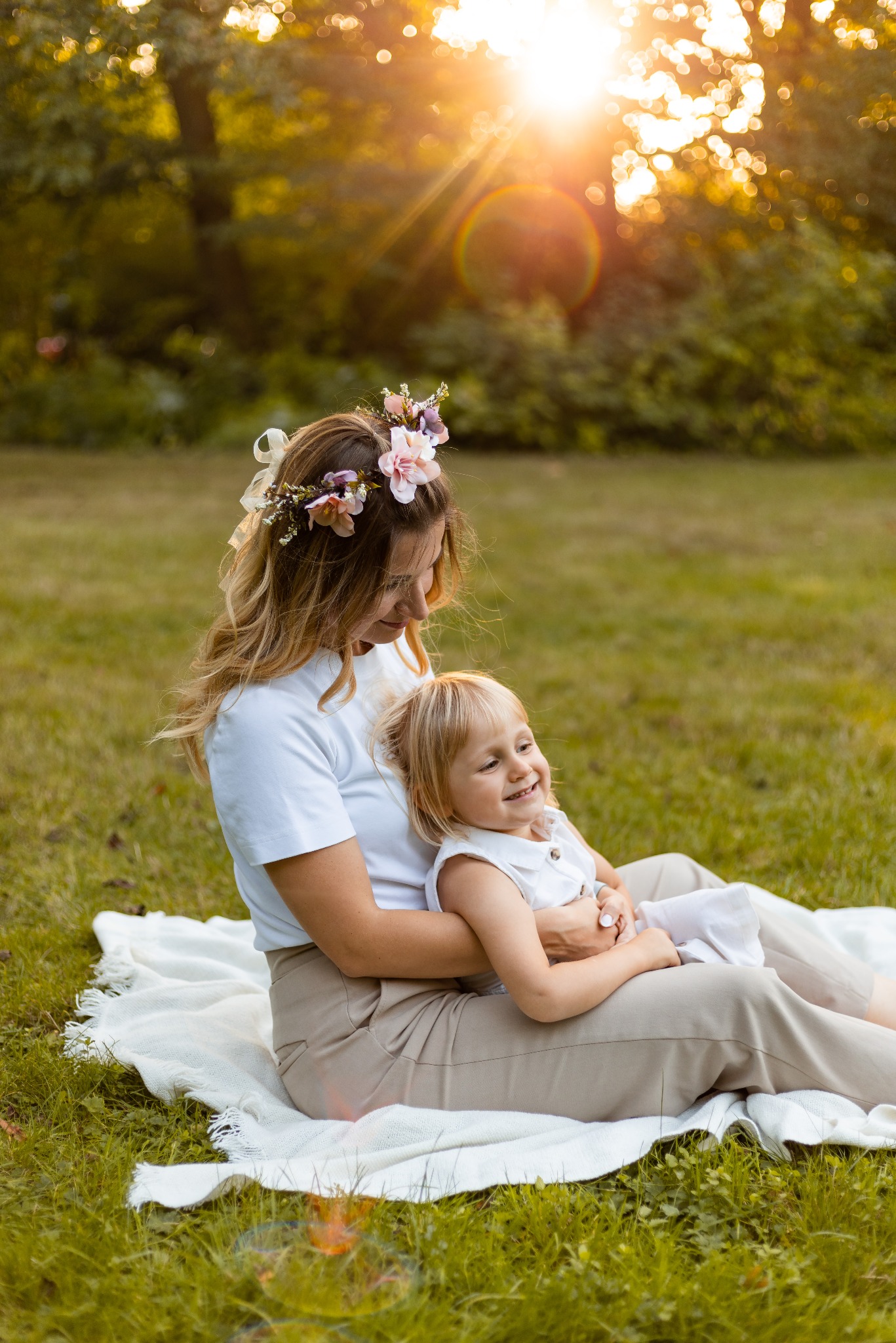 Mama z córeczką w wianku siedzą na kocu w trawie, w tle słońce. Jasne ubrania, naturalne światło, ciepła atmosfera. Sesja rodzinna w plenerze.