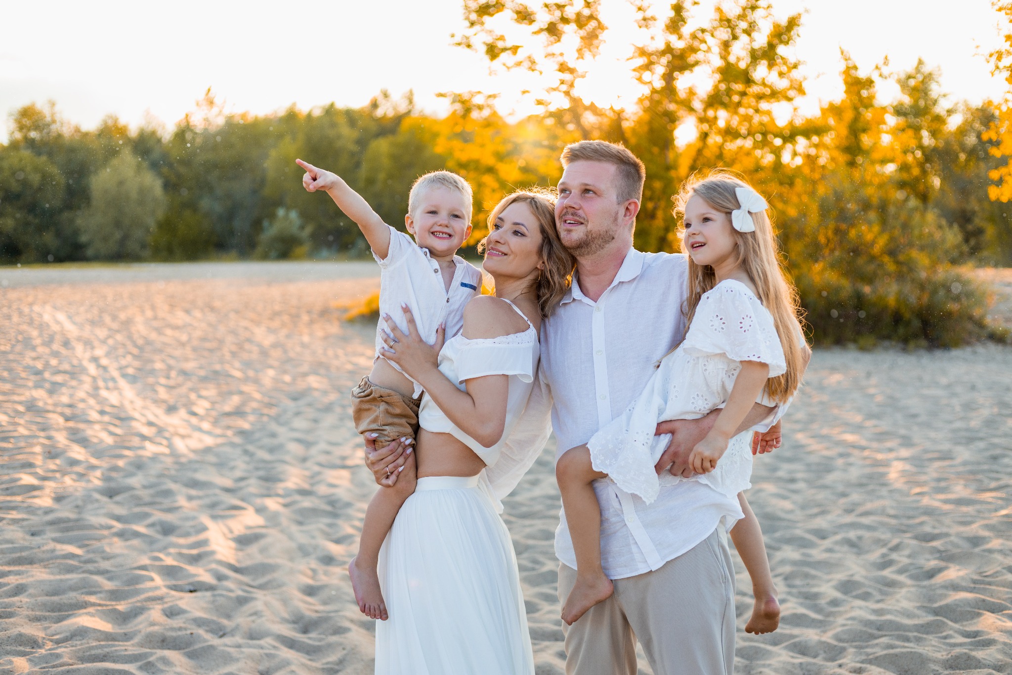 Rodzinna sesja w Warszawie: Mama trzyma synka wskazującego palcem w górę, tata córeczkę, wszyscy uśmiechnięci na tle piaszczystej plaży i drzew w ciepłym świetle.