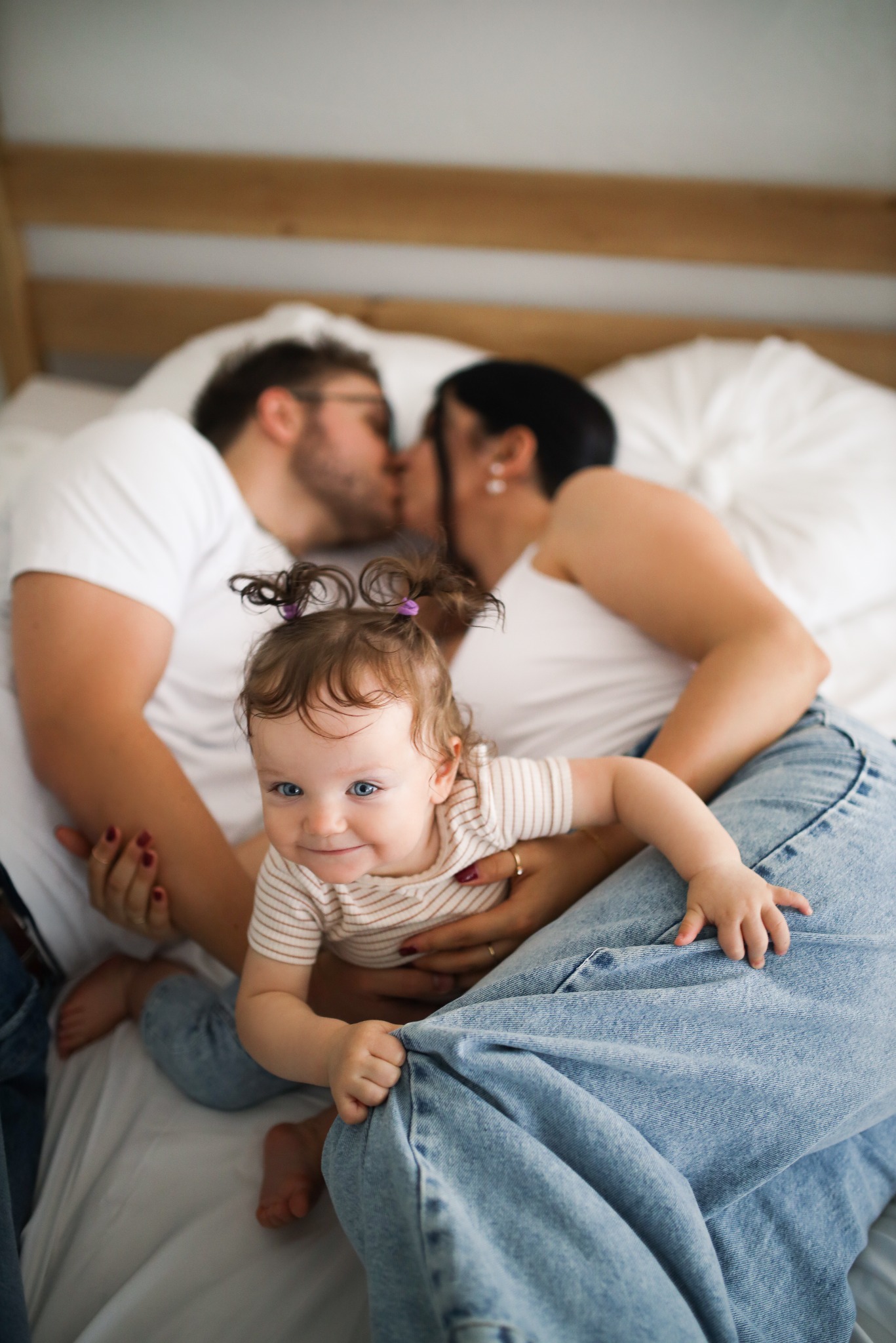 Rodzinna sesja w łóżku: uśmiechnięta dziewczynka na kolanach mamy, w tle całująca się para. Jasne, naturalne światło, miękka pościel, dżinsowe spodnie.