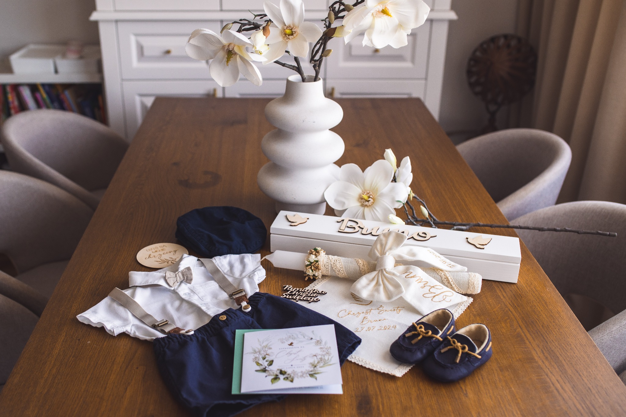 Kompozycja z ubrankami, świecą i zaproszeniem na chrzest na drewnianym stole, z dekoracyjną wazą z magnoliami w tle. Elementy ceremonii chrztu.