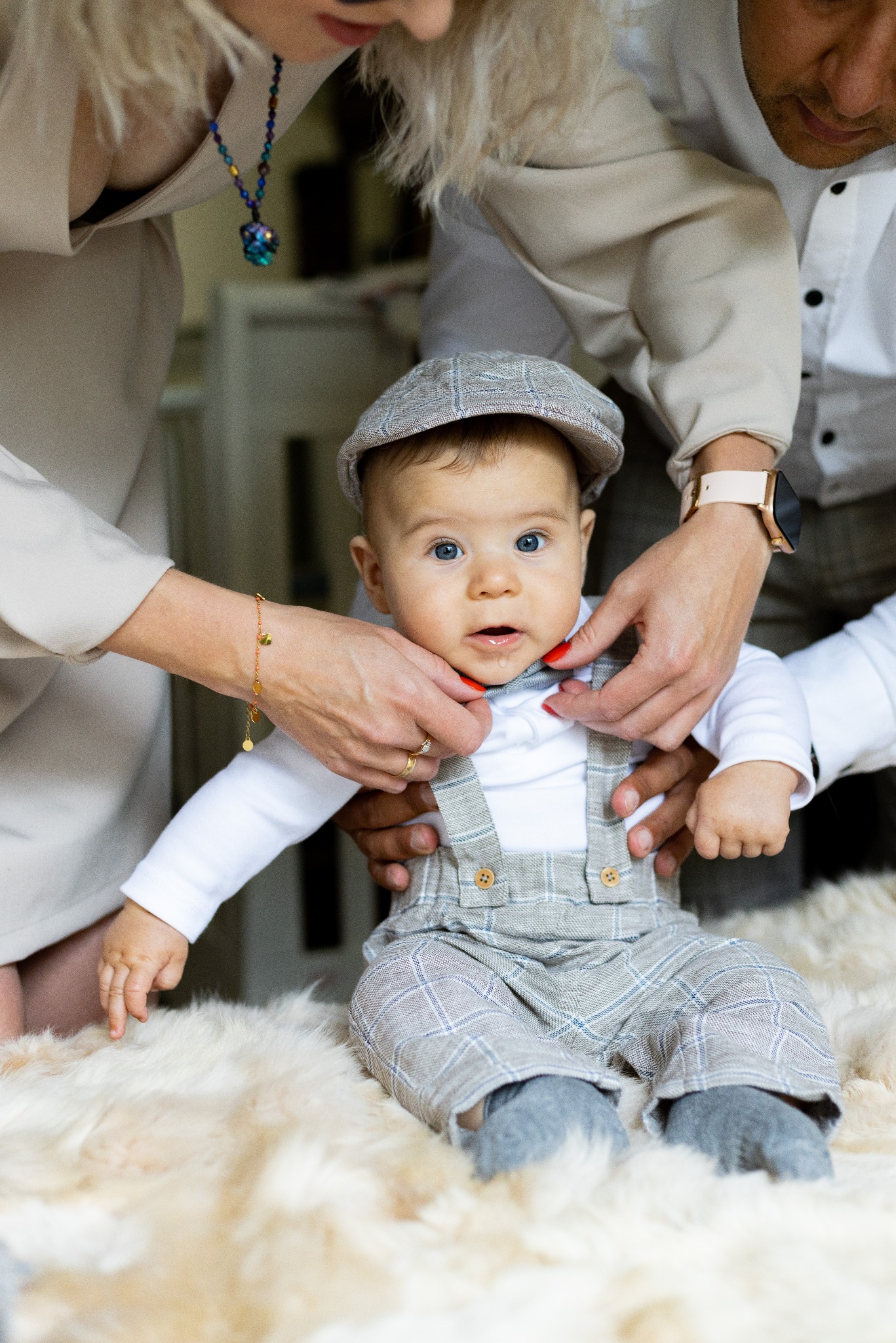 Uroczy niemowlak w kraciastej czapce i spodniach na futrzanym dywanie, poprawiany przez rodziców. Jasne, naturalne światło podkreśla dziecięcą niewinność.