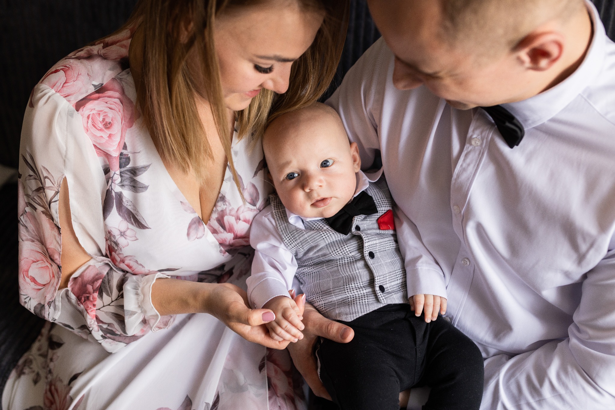 Rodzinna fotografia: Mama w kwiecistej sukience i tata w koszuli z muchą trzymają elegancko ubranego synka w kamizelce i muszce. Dziecko patrzy w obiektyw.