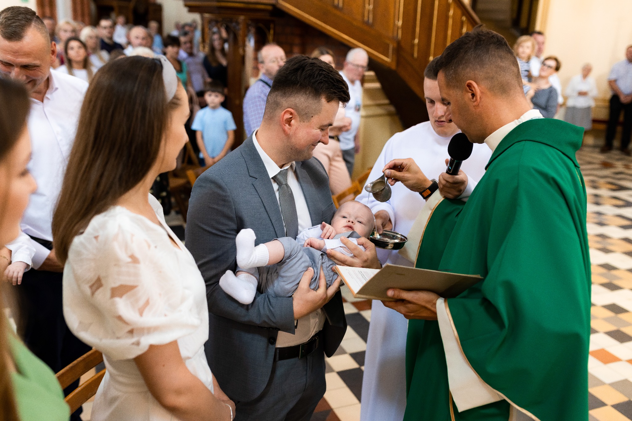 Chrzest dziecka w kościele: ojciec trzyma niemowlę, ksiądz polewa wodą z naczynia. W tle rodzina i goście, wnętrze kościoła z drewnianymi schodami.