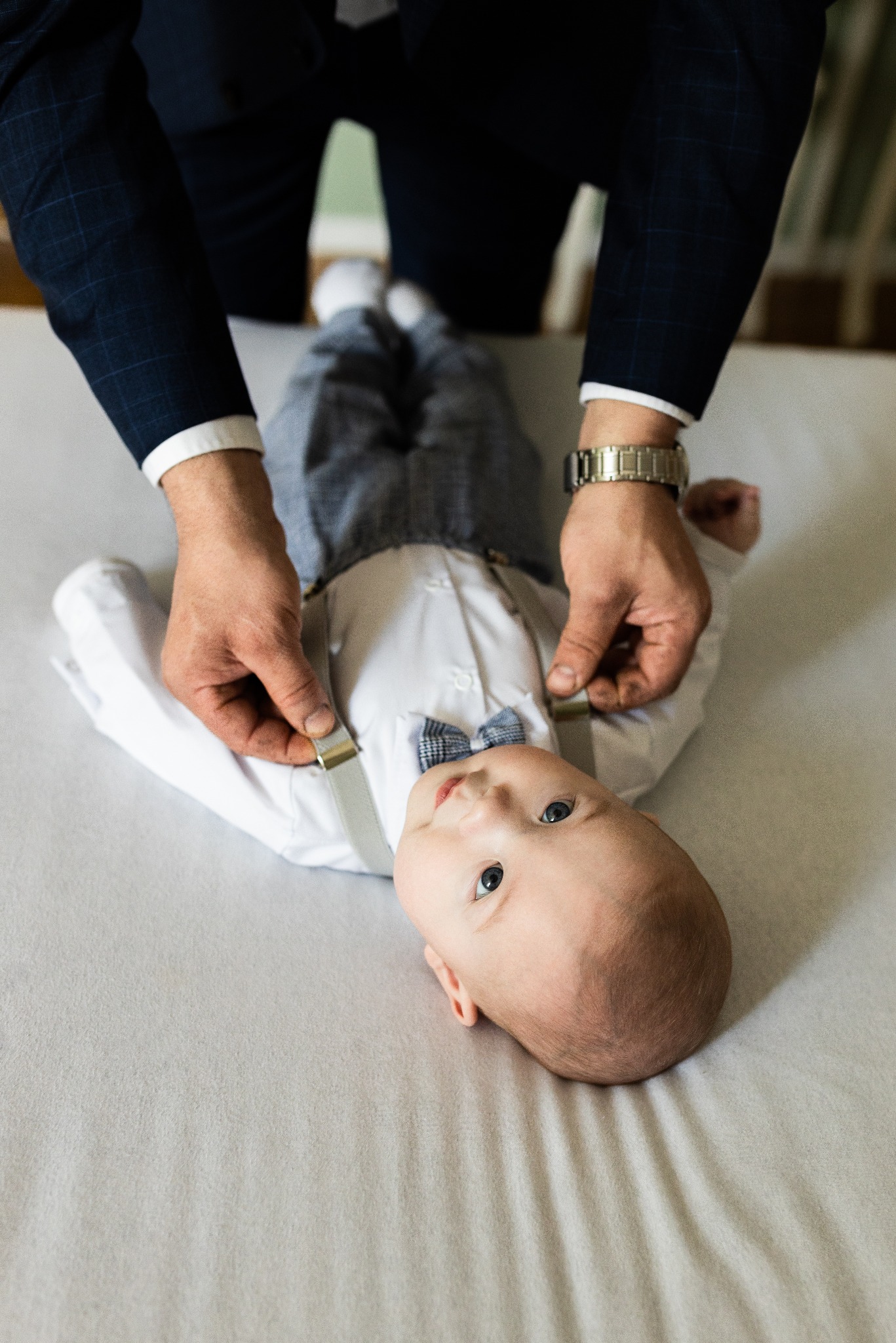 Uśmiechnięte niemowlę w eleganckim stroju z muchą i szelkami, poprawianymi przez dorosłego w garniturze, leżące na jasnym tle. Ujęcie z góry, naturalne światło.